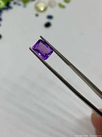 Amethyst gemstone held by tweezers showing rectangular cushion shape and purple hue