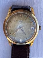 Close-up of watch face showing brand, dial patina, gold indices, date window, crown, and case material