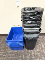 Stack of 5 blue recycling bins beside stack of black and grey trash cans with liners