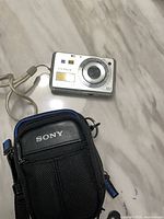 Front view of silver Sony Cyber-shot camera with wrist strap beside black Sony case