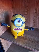 Front view of yellow one-eyed Minion plastic figure with arms outstretched on wooden shelf