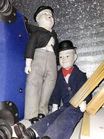 Both dolls seated/upright showing outfits, hats, porcelain heads and hands