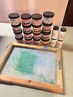 Stacked jars of Speedball screen printing ink, auxiliary fluid bottles, wood frame screen