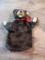 Front view of brown bear hand puppet lying flat showing red ribbon, beige felt accents