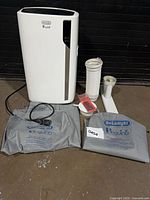 Front view of DeLonghi Pinguino portable air conditioner with accessories and storage bags laid out