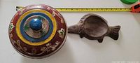 Lidded round painted bowl beside carved fish bowl with measuring tape for scale