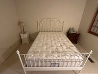 Overall view of white metal queen bed frame, white nightstand with lamp, dark wood nightstand with fax machine and phone