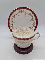 Cup positioned before upright saucer showing burgundy border and gold vine design