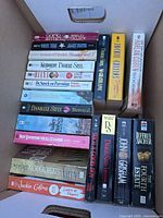 Top-view of box showing two stacks of books with visible titles by Nora Roberts, Jackie Collins, Danielle Steel, Harlan Coben, Jeffrey Archer and others.