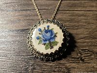 Front view of pendant showing blue rose needlepoint and scalloped metal frame