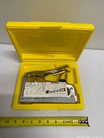 Staple gun inside open yellow case with instruction sheet, tape measure showing size