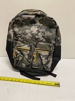 Front view showing camo skull pattern, two front pockets, yellow zipper pulls, measuring tape for scale