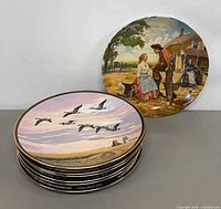 Stack of seven gold-rimmed collector plates, top plate shows geese in flight, one plate displayed upright