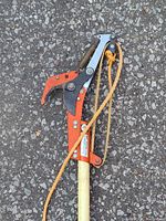 Close view of orange metal lopper head, spring, blade and pull rope on light yellow pole