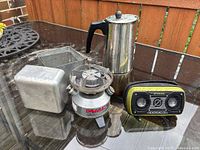 Coleman stove, aluminum case, stainless percolator and Rock Out 2 speaker displayed together