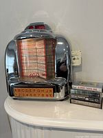 Front angled view of chrome tabletop jukebox with stack of music cases beside it