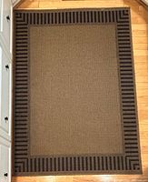 Full view of rectangular brown rug with black striped border on hardwood floor