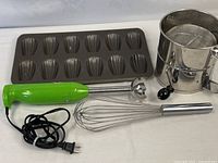 Four items laid out: immersion blender, whisk, Madeleine pan, flour sifter
