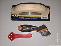 Top view of gum rubber grout float in packaging, RIDGID grout saw, red caulk applicator