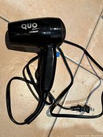 Full view of black Quo Beauty by Conair compact travel hair dryer with cord and plug on tile