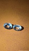 Two light blue oval faceted aquamarines on brown background, face-up view