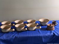 All pottery pieces on blue cloth including six handled bowls, two regular bowls, brown mug, brown lidded crock