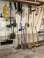 Overall view of hanging and leaning yard tools including snow shovel, shovels, brooms, weed trimmers, cords, pole pruner