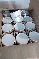 Open box containing grid-divided 24 white mugs, one logo side visible