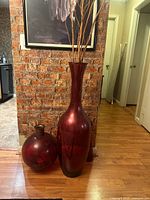 Tall red vase with branches beside round red vase against brick wall