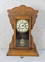 Front view of carved oak mantel clock showing full case, dial, glass door design, pendulum and key