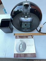 Front top view of black Nescafe Dolce Gusto coffee machine, power cord, capsule bin and ruler for scale, manual visible