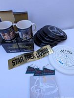 Group shot showing mugs in boxes, stack of black frisbees, white frisbee, bumper sticker, key chains on cards, and notepad