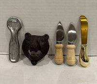 All five items lined up: handheld bottle opener, bear-head wall-mount bottle opener, two wood-handled cheese knives, gold-tone spreader