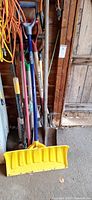 Group of long-handled tools leaning against wall showing snow shovel, broom, spade, fork and rake heads