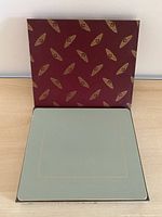 Open box showing sage green placemat with gold border and patterned lid