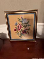 Framed needlepoint of floral bouquet with butterfly in gold frame and blue mat behind glass