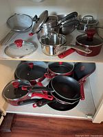 All cookware pieces arranged on two cabinet shelves showing red nonstick pans, stainless pans, strainer, and lids