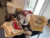 Wide view of all items on table: pots, kettles, dish rack with oven mitts, spice rack, cake stand, mixer parts in tub