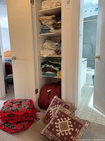 Open linen closet shelves filled with sheets, throws, red duffle, cushions on floor