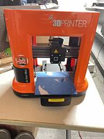 Front view showing orange body, print head, print bed with blue tape