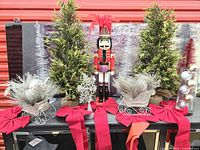 Front view of nutcracker flanked by three evergreen trees, silver sleighs, bows, wire trees and vase
