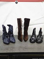Front view of three pairs of boots on table: navy wrap booties, brown suede tall boots, black leather ankle boots