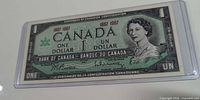 Obverse of 1967 Canada $1 bill in sleeve showing Queen Elizabeth II portrait, denomination, centennial dates, clear signatures