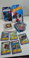 Front of sealed Hot Wheels Snoopy car, G.I. Joe figure, stacks of Digimon and Exploding Kittens cards