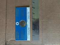 Coin in blue presentation sleeve beside ruler for scale