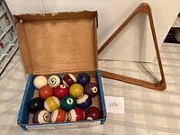 Box holding 15 colored billiard balls plus cue ball next to wooden triangle rack
