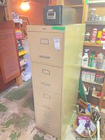 Front view of beige four-drawer metal filing cabinet with black index card box on top, surrounding shelves visible