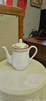 White porcelain teapot with gold trim and lid displayed on lace surface
