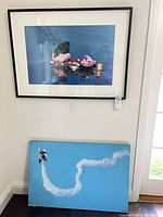 Both artworks leaning against wall: framed Robert Barab print above, panda rocket canvas below