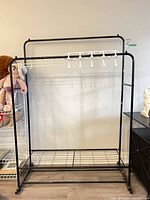 Full view of black metal garment rack with top and mid bars, bottom wire shelf, partial view of surrounding items